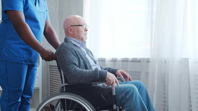 African-American caregiver and old disabled man in a wheelchair. Professional nurse and handicapped patient in a nursing home. Assistance, rehabilitation and health care.