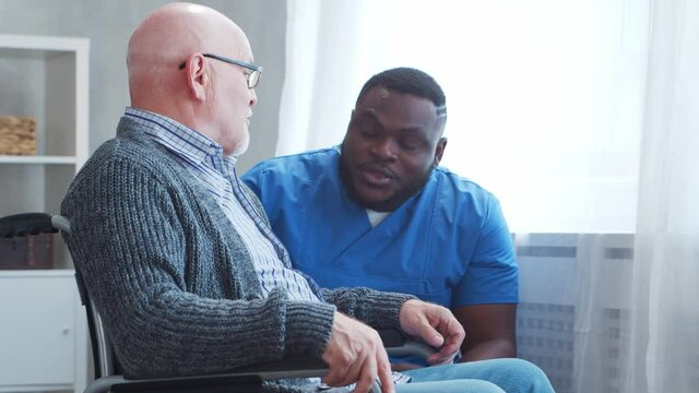 African-American caregiver and old disabled man in a wheelchair. Professional nurse and handicapped patient in a nursing home. Assistance, rehabilitation and health care.