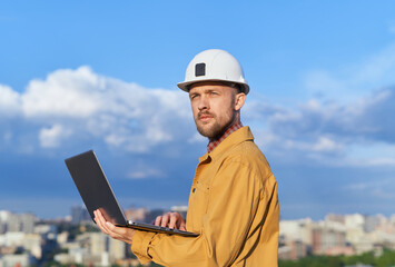 Attractive caucasian bearded engineer using laptop at construction site wearing orange jacket and hard hat as protective workwear. Sunny day with blue cloudy sky and urban skyline. High quality photo