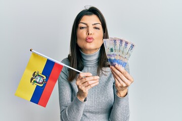 Young hispanic woman holding colombia flag and colombian pesos banknotes looking at the camera blowing a kiss being lovely and sexy. love expression.