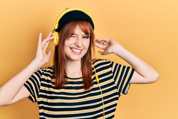 Redhead young woman listening to music using headphones winking looking at the camera with sexy expression, cheerful and happy face.