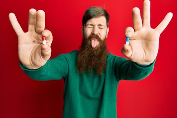 Redhead man with long beard holding two different pills sticking tongue out happy with funny...