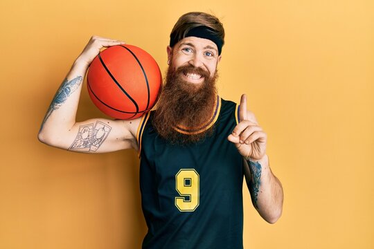 Redhead man with long beard wearing basketball uniform holding ball smiling with an idea or question pointing finger with happy face, number one