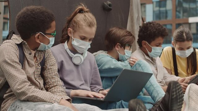 Slowmo shot of group of multiethnic preteens in face masks spending time outdoors after school using laptop and digital tablet while doing home assignment