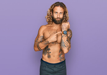 Handsome man with beard and long hair standing shirtless showing tattoos in hurry pointing to watch...