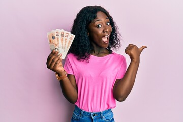 Beautiful african young woman holding 10 united kingdom pounds banknotes pointing thumb up to the side smiling happy with open mouth