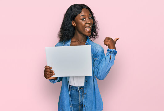 Beautiful african young woman holding blank empty banner pointing thumb up to the side smiling happy with open mouth
