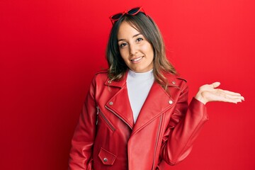 Young brunette woman wearing red leather jacket smiling cheerful presenting and pointing with palm of hand looking at the camera.