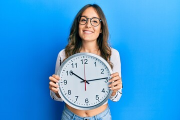 Young brunette woman holding big clock smiling with a happy and cool smile on face. showing teeth.