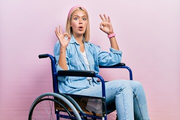 Beautiful blonde woman sitting on wheelchair looking surprised and shocked doing ok approval symbol...