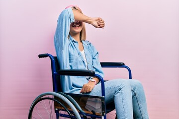 Beautiful blonde woman sitting on wheelchair covering eyes with arm smiling cheerful and funny....