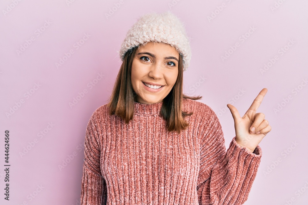 Wall mural Young beautiful woman wearing wool sweater and winter hat smiling cheerful pointing with hand and finger up to the side