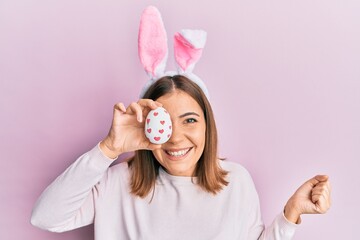 Young beautiful woman wearing cute easter bunny ears holding egg screaming proud, celebrating victory and success very excited with raised arm