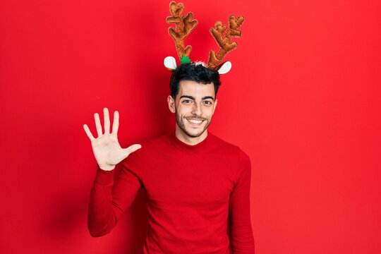 Young hispanic man wearing cute christmas reindeer horns showing and pointing up with fingers number five while smiling confident and happy.