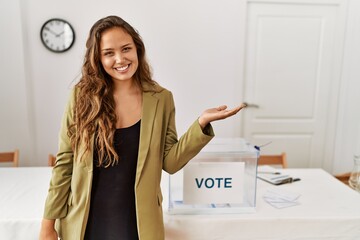 Beautiful hispanic woman standing at political campaign room smiling cheerful presenting and pointing with palm of hand looking at the camera.