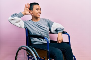 Beautiful hispanic woman with short hair sitting on wheelchair smiling with hand over ear listening...