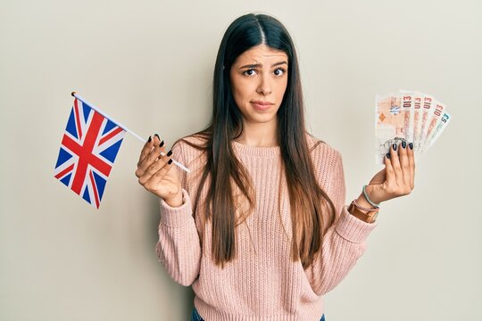 Young hispanic woman holding united kingdom flag and pounds banknotes skeptic and nervous, frowning upset because of problem. negative person.