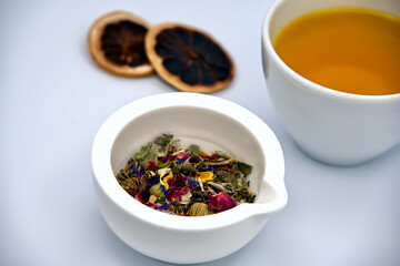Dried herbs and flower petals in a white mortar, a cup of fresh tea and dried orange slices against a white background
