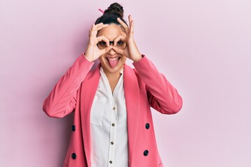 Beautiful middle eastern woman wearing business jacket and glasses doing ok gesture like binoculars sticking tongue out, eyes looking through fingers. crazy expression.