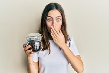 Young brunette woman holding jar with coffee beans covering mouth with hand, shocked and afraid for...