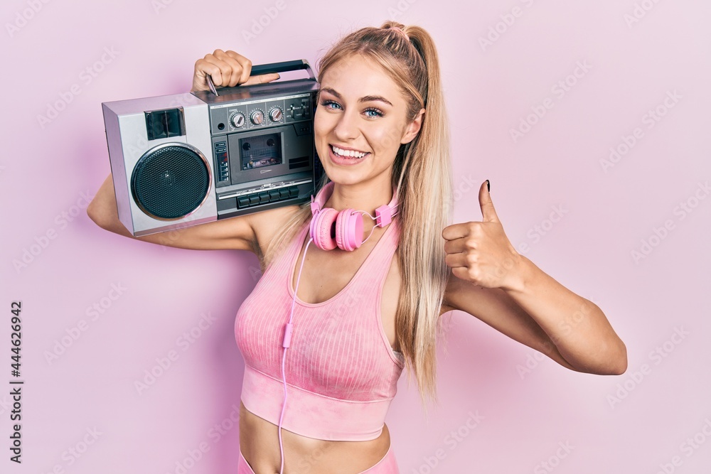 Wall mural Young beautiful caucasian woman wearing workout clothes and holding boombox smiling happy and positive, thumb up doing excellent and approval sign