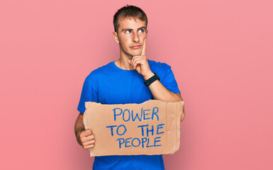 Young blond man holding power to the people banner serious face thinking about question with hand...
