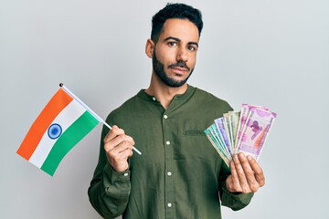 Young hispanic man holding india flag and rupee banknotes puffing cheeks with funny face. mouth inflated with air, catching air.
