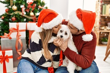 Young hispanic couple wearing christmas hat sitting on the floor with dog at home.
