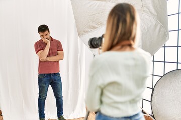 Woman photographer talking pictures of man posing as model at photography studio thinking looking tired and bored with depression problems with crossed arms.