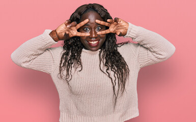 Young african woman wearing wool winter sweater doing peace symbol with fingers over face, smiling cheerful showing victory