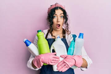 Young brunette woman with curly hair wearing cleaner apron holding cleaning products afraid and shocked with surprise and amazed expression, fear and excited face.