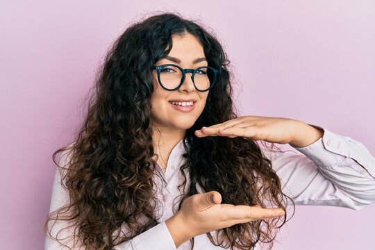 Young brunette woman with curly hair wearing casual clothes and glasses gesturing with hands showing big and large size sign, measure symbol. smiling looking at the camera. measuring concept.