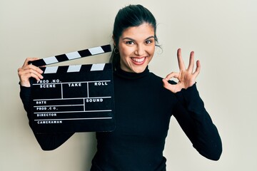 Young hispanic woman holding video film clapboard doing ok sign with fingers, smiling friendly...