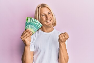 Caucasian young man with long hair holding 20000 indonesian rupiah screaming proud, celebrating victory and success very excited with raised arm