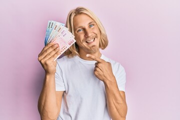 Caucasian young man with long hair holding new taiwan dollars banknotes smiling happy pointing with hand and finger