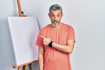 Handsome middle age man with grey hair standing by painter easel stand surprised pointing with finger to the side, open mouth amazed expression.