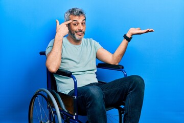 Handsome middle age man with grey hair sitting on wheelchair confused and annoyed with open palm showing copy space and pointing finger to forehead. think about it.