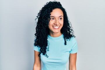 Young hispanic woman with curly hair wearing casual blue t shirt looking away to side with smile on face, natural expression. laughing confident.