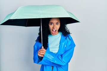 Young hispanic woman with curly hair wearing a raincoat and umbrella celebrating crazy and amazed for success with open eyes screaming excited.