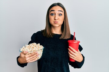 Young brunette girl eating popcorn and drinking soda puffing cheeks with funny face. mouth inflated...