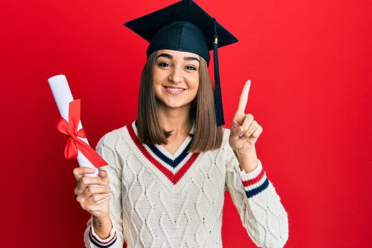 Young brunette girl holding graduate degree diploma smiling with an idea or question pointing finger with happy face, number one
