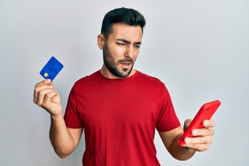 Young hispanic man holding smartphone and credit card clueless and confused expression. doubt...