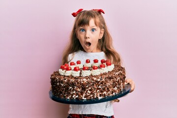 Little caucasian girl kid celebrating birthday holding big chocolate cake afraid and shocked with...