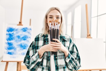 Young artist student girl smiling happy holding paintbrushes covering face at art studio.