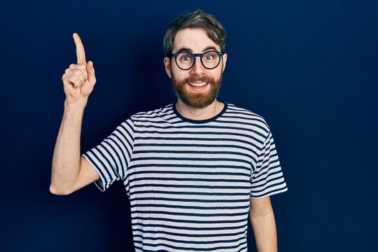 Caucasian man with beard wearing striped t shirt and glasses pointing finger up with successful idea. exited and happy. number one.