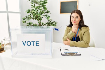 Young brunette woman sitting at election table with voting ballot looking confident at the camera...