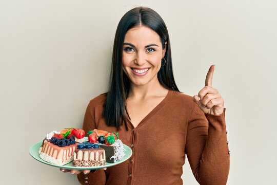 Young brunette woman holding plate with cake slices smiling with an idea or question pointing finger with happy face, number one