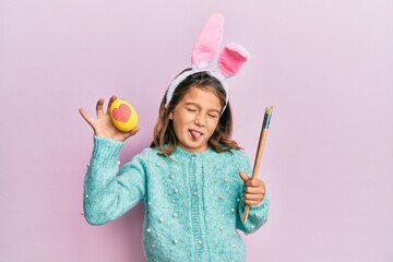 Little beautiful girl wearing cute easter bunny ears holding colored egg sticking tongue out happy...