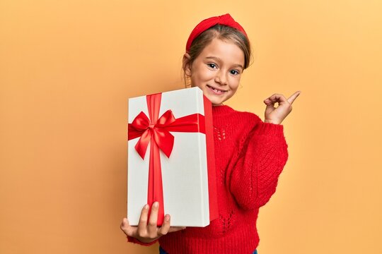 Little beautiful girl holding gift smiling happy pointing with hand and finger to the side