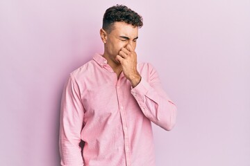 Hispanic young man wearing casual clothes smelling something stinky and disgusting, intolerable smell, holding breath with fingers on nose. bad smell